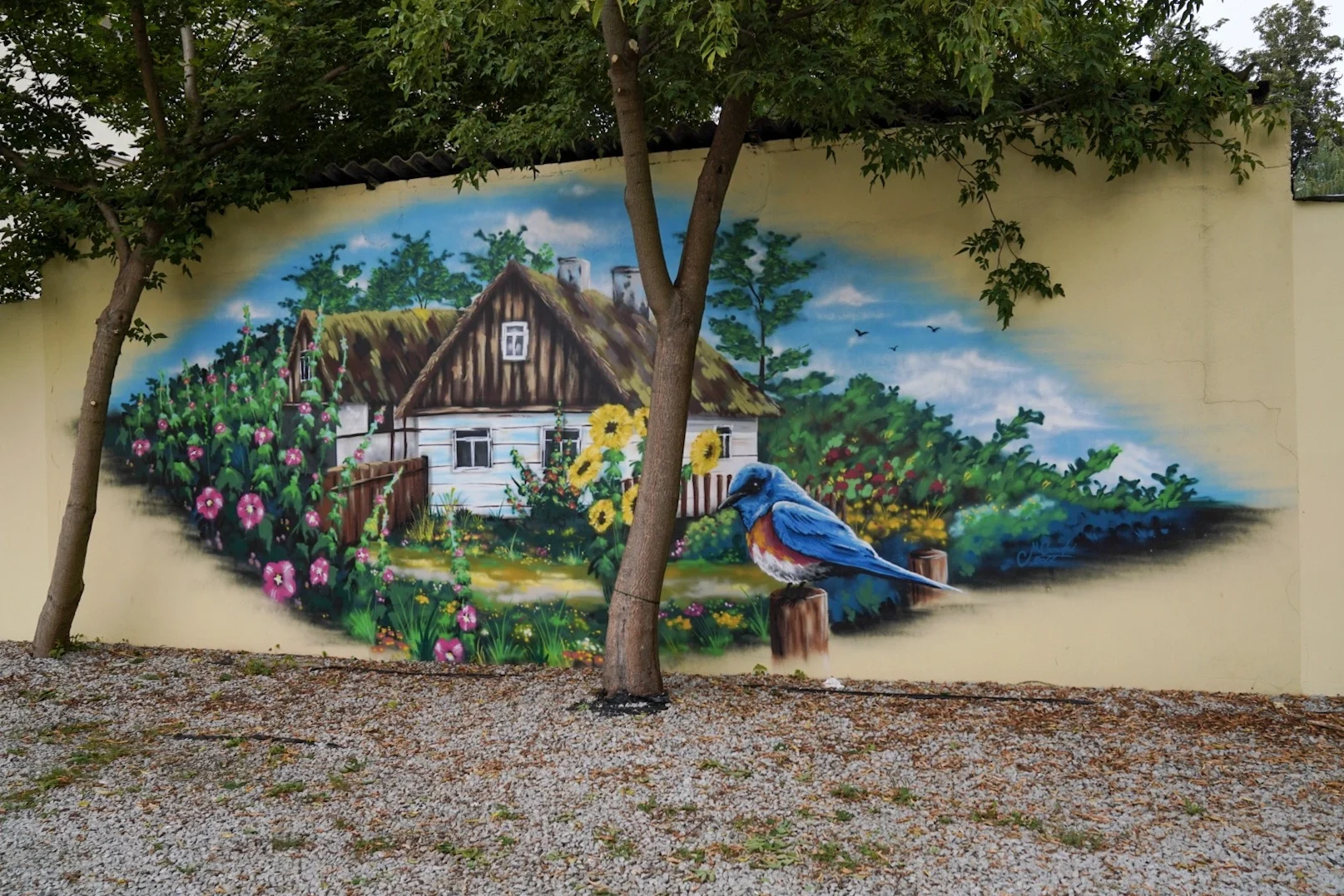 Kolorowy mural w Białymstoku przedstawiający drewniany dom, ogród pełen kwiatów i niebieskiego ptaka na tle wiejskiego krajobrazu.