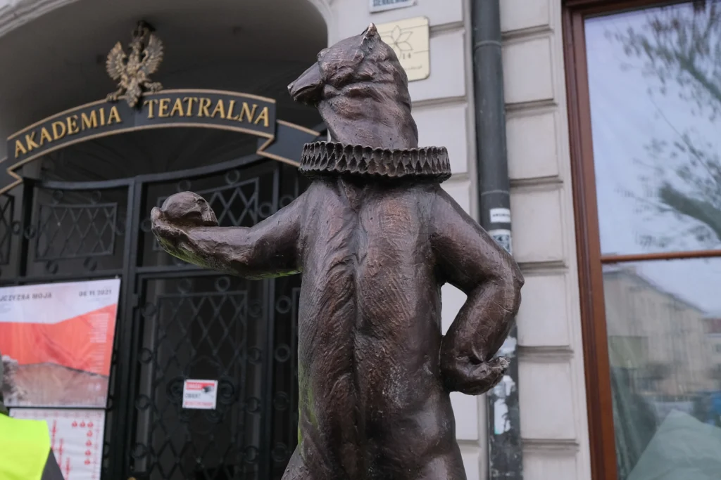 igurka białostockiego WidziMisia w teatralnym kołnierzu stojąca przed wejściem do Akademii Teatralnej w Białymstoku.
